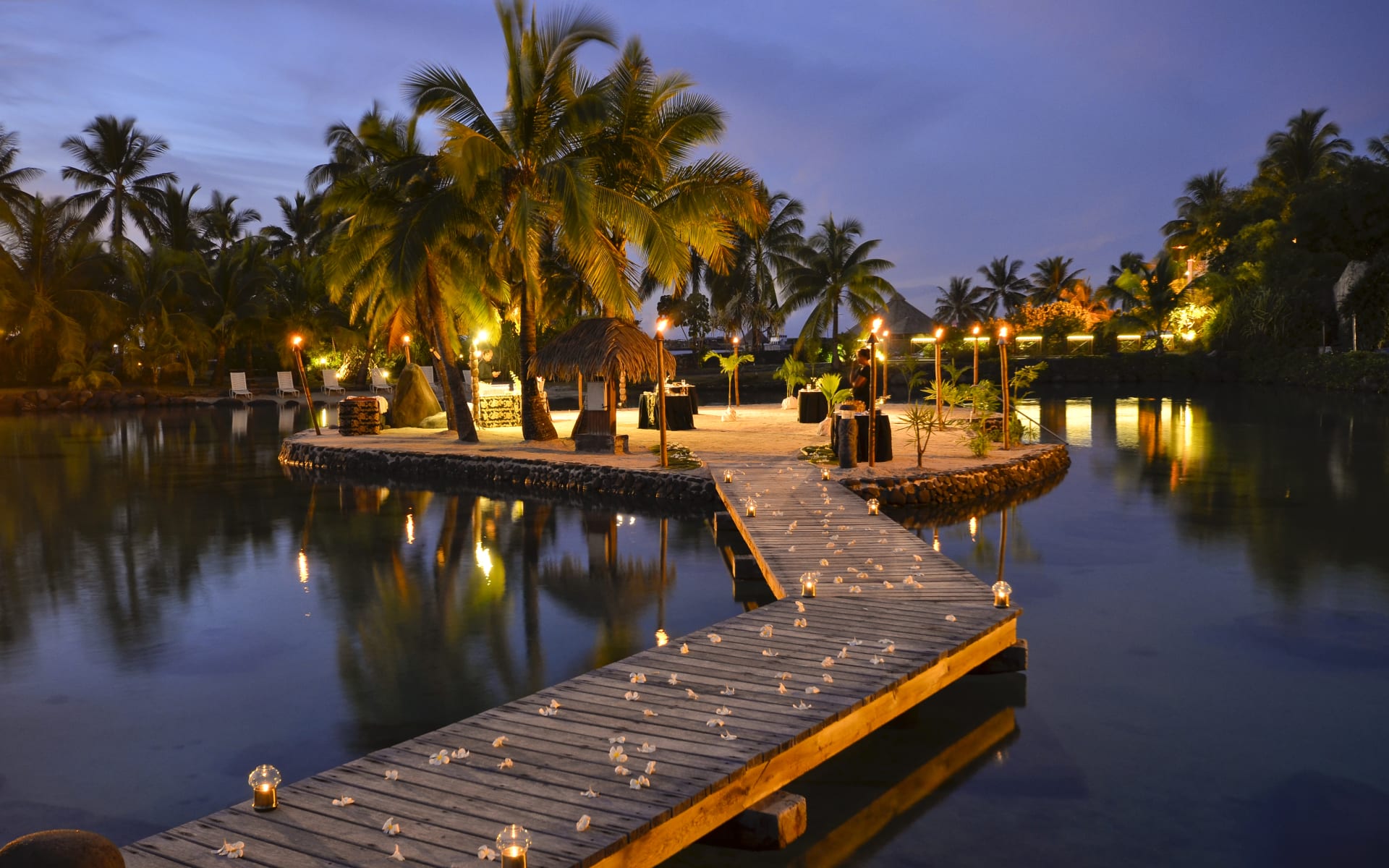 A wooden pathway leads to an island with chairs, lights and tables. 