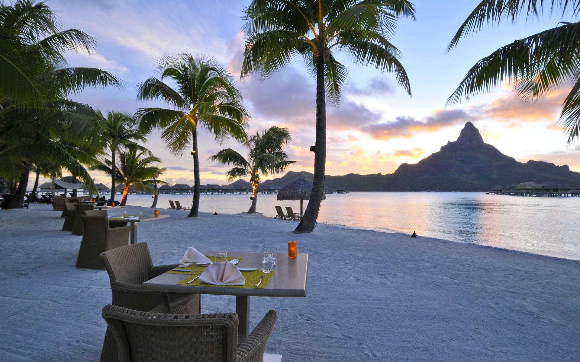 Two-person tables are lined on the beach, overlooking the ocean, mountains and palm trees. 