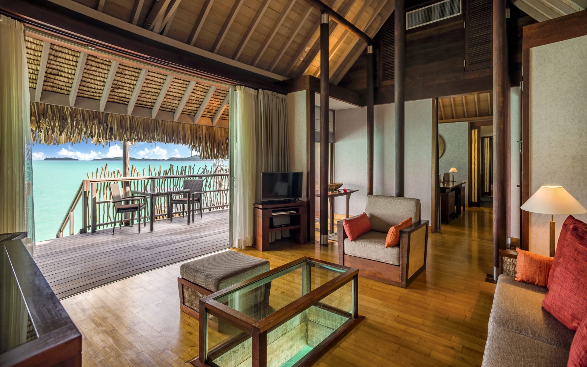The living room has sofas, a table and a TV with floor-to-ceiling doors leading to the deck and ocean. 