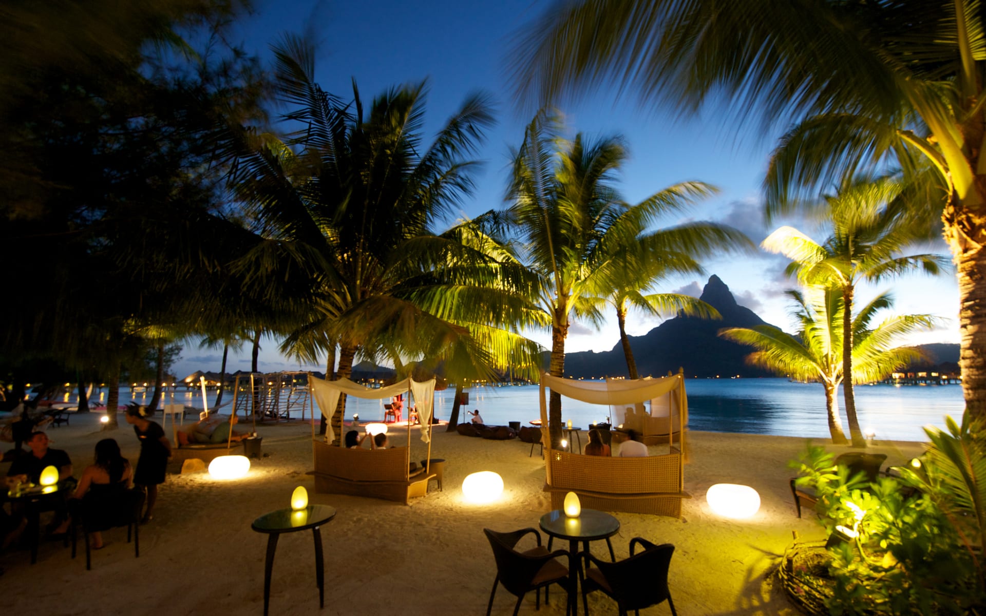 The beach has palm trees, tables, cabanas and lantern lighting. 