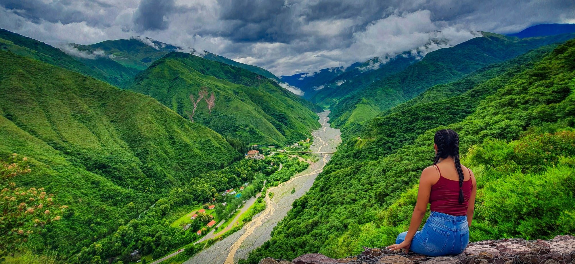 A woman in a red top is sitting on a stone wall overlooking the green, vibrant v-shaped valley in Jujuy. 