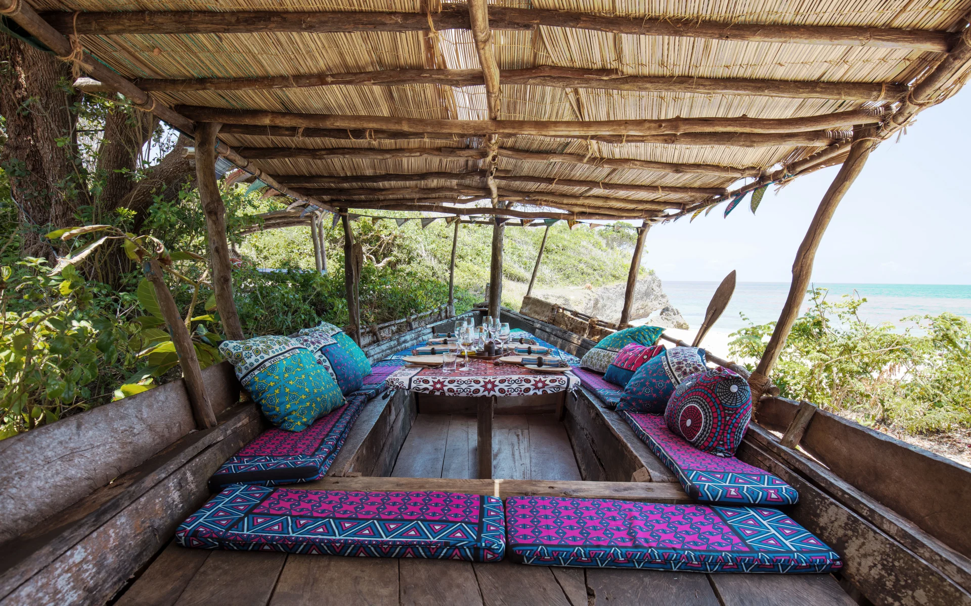A wooden boat is dressed with cosy, patterned seating and a dining spread. It is shaded and on the shore of Galu Beach.