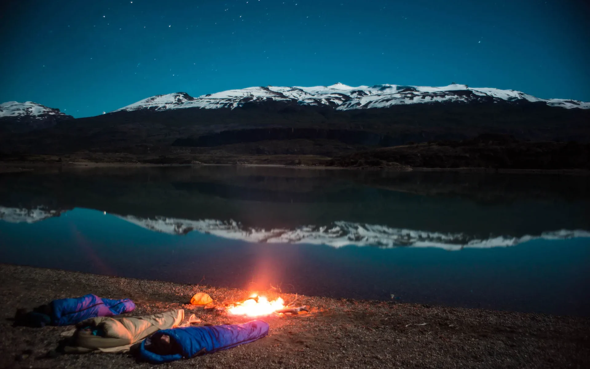 Three people cosy up in sleeping bags beside a log fire, ahead of an evening scene of the lake and the tumbling Andes.