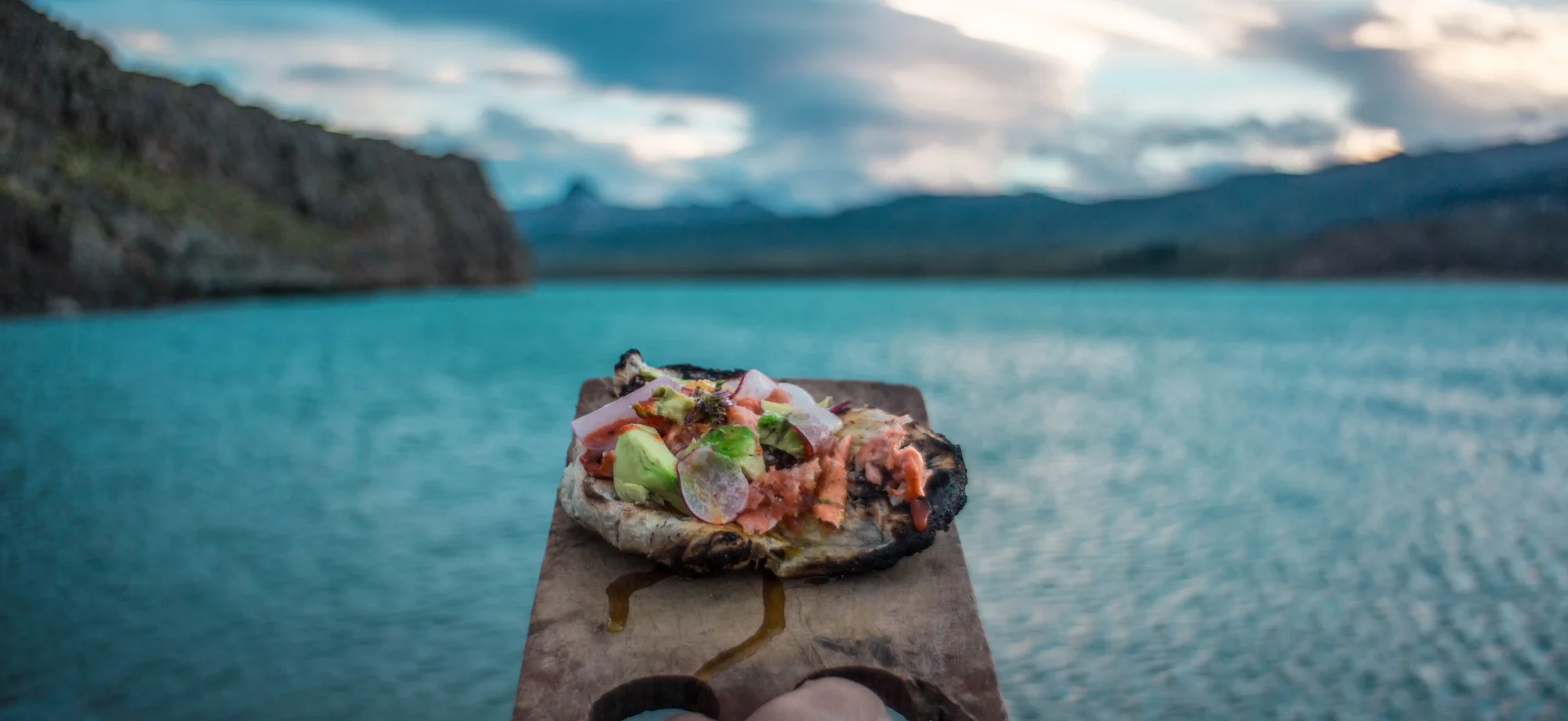 Someone is holding a delicious traditional dish, served on a slab of wood, ahead of a stunning scene of Lago Argentino.