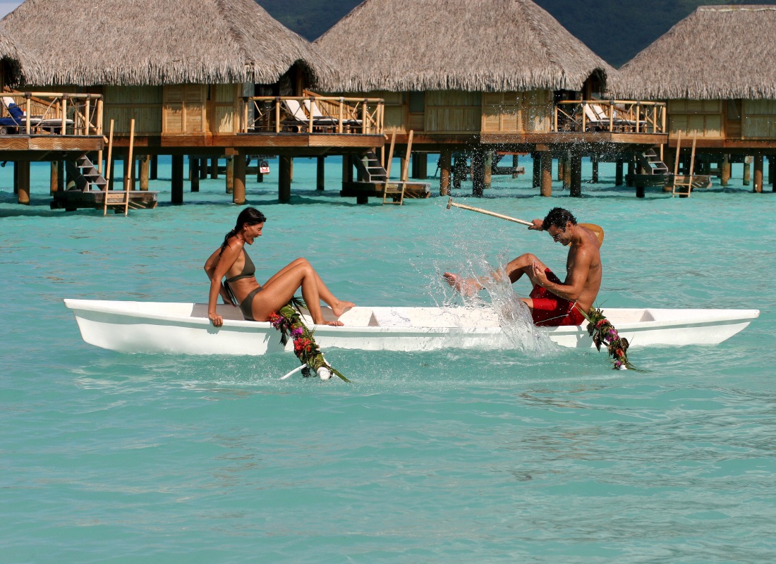 A couple are sitting on a boat next to thatched-roof water villas. 
