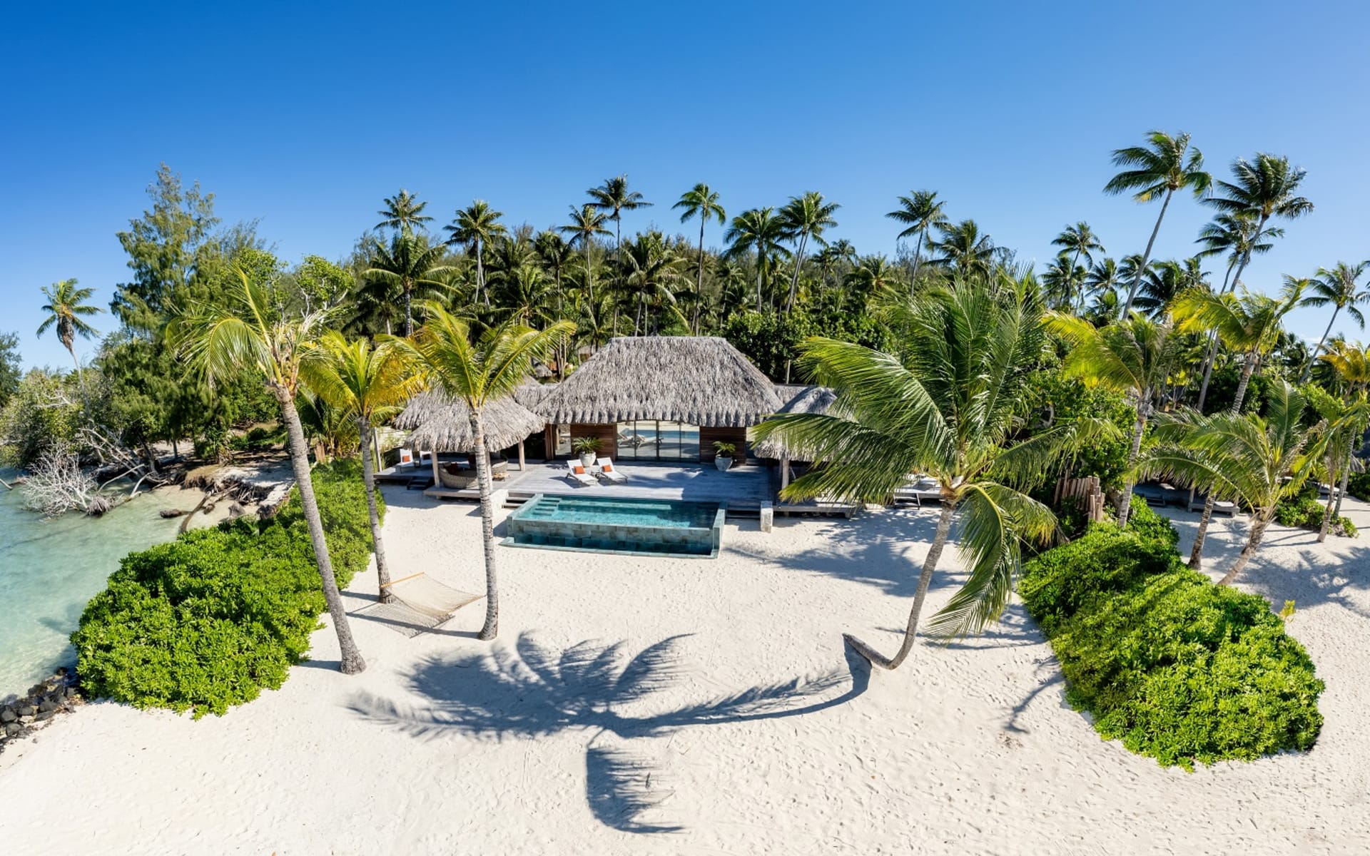 A massive thatched-roof villa with a private pool opens onto the beach and is surrounded by palm trees. 