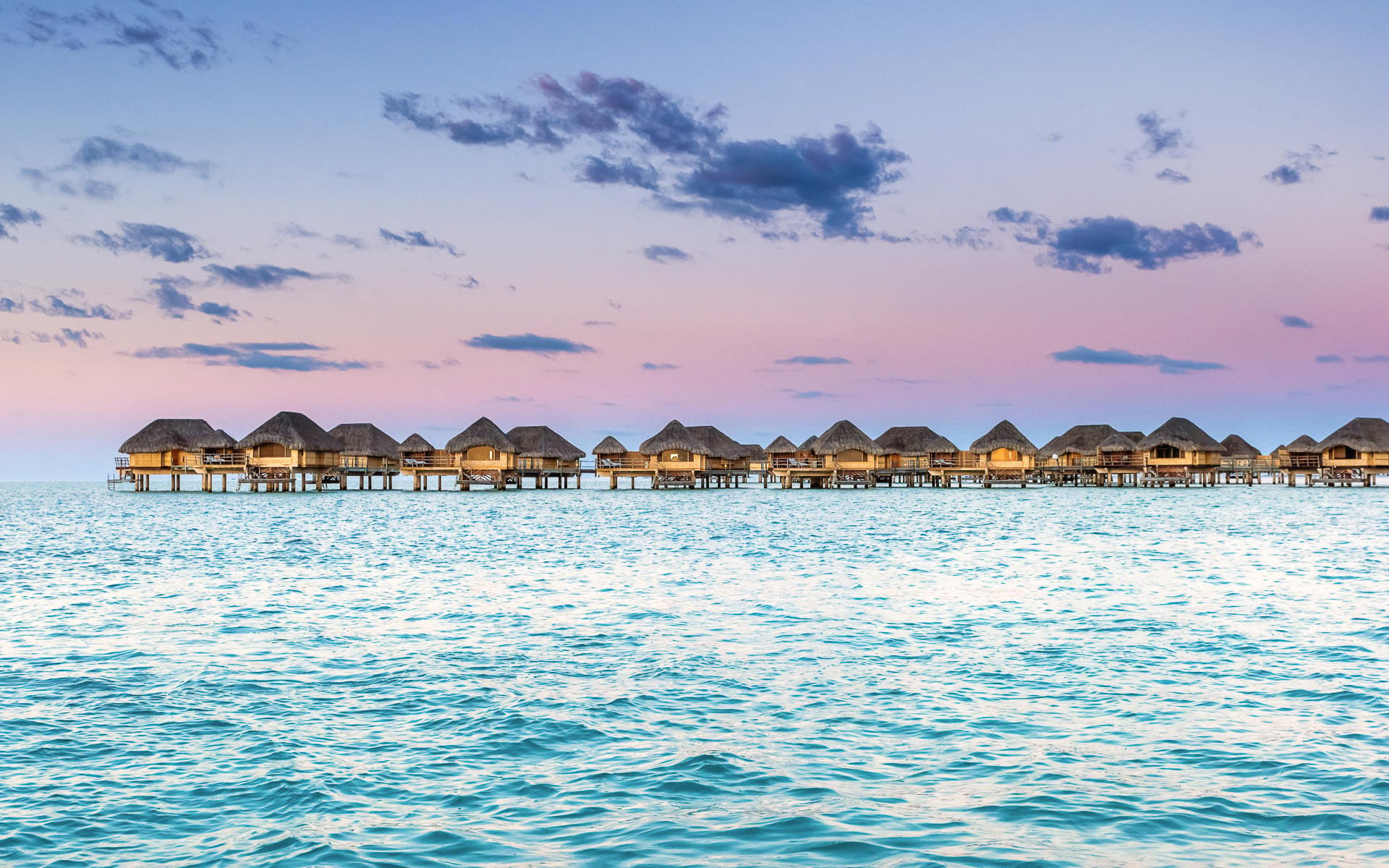 There is a row of overwater villas backed by a purple-blue sunset. 