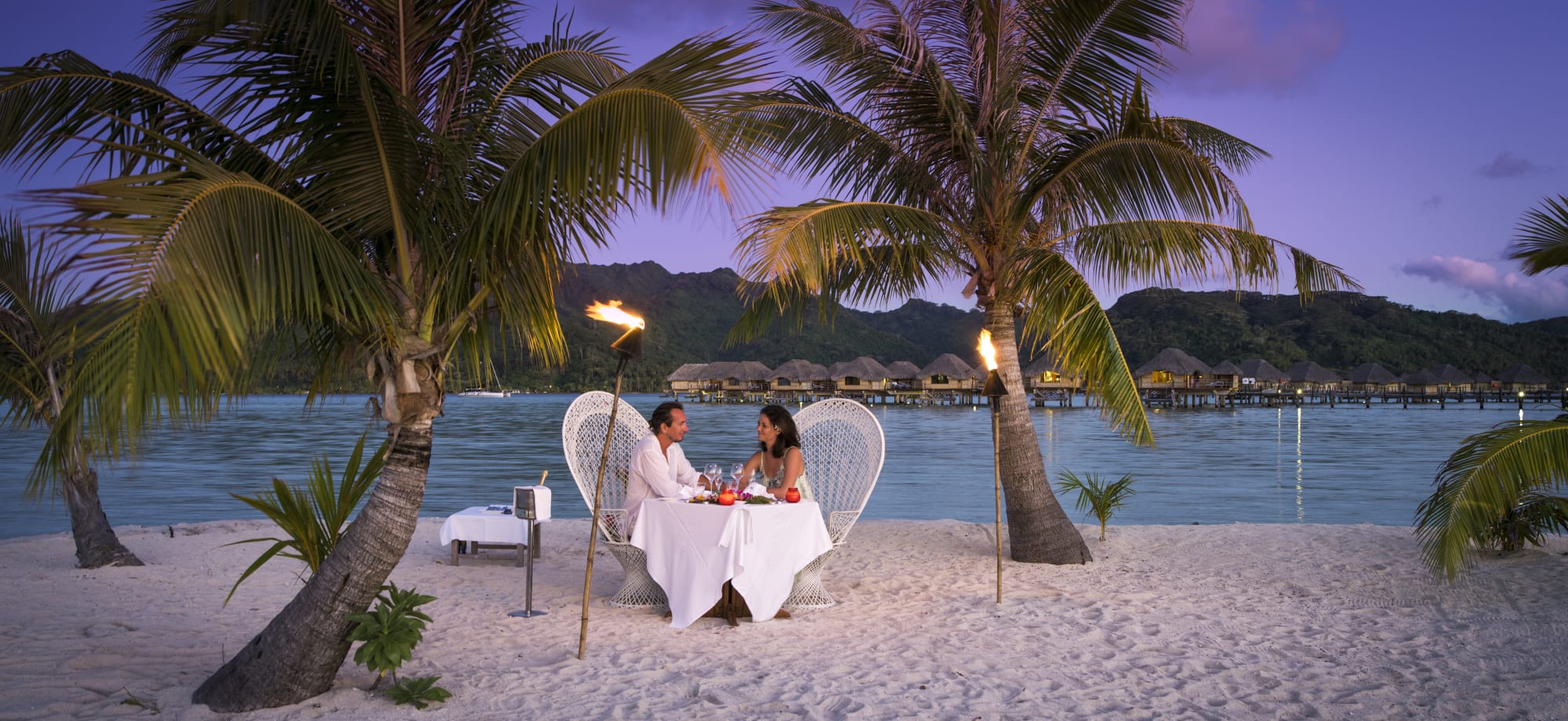 A couple are sitting at a table on the beach as the sun sets. 