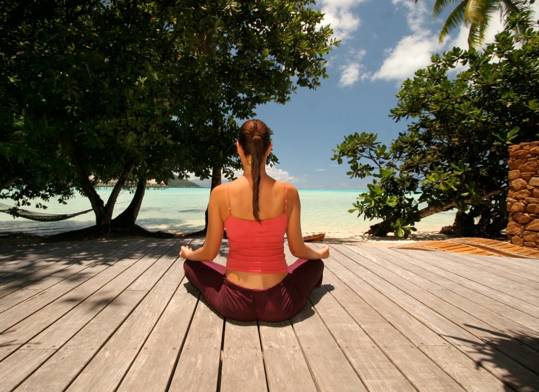 A woman is sitting on a wooden deck overlooking the ocean and doing meditation and yoga. 