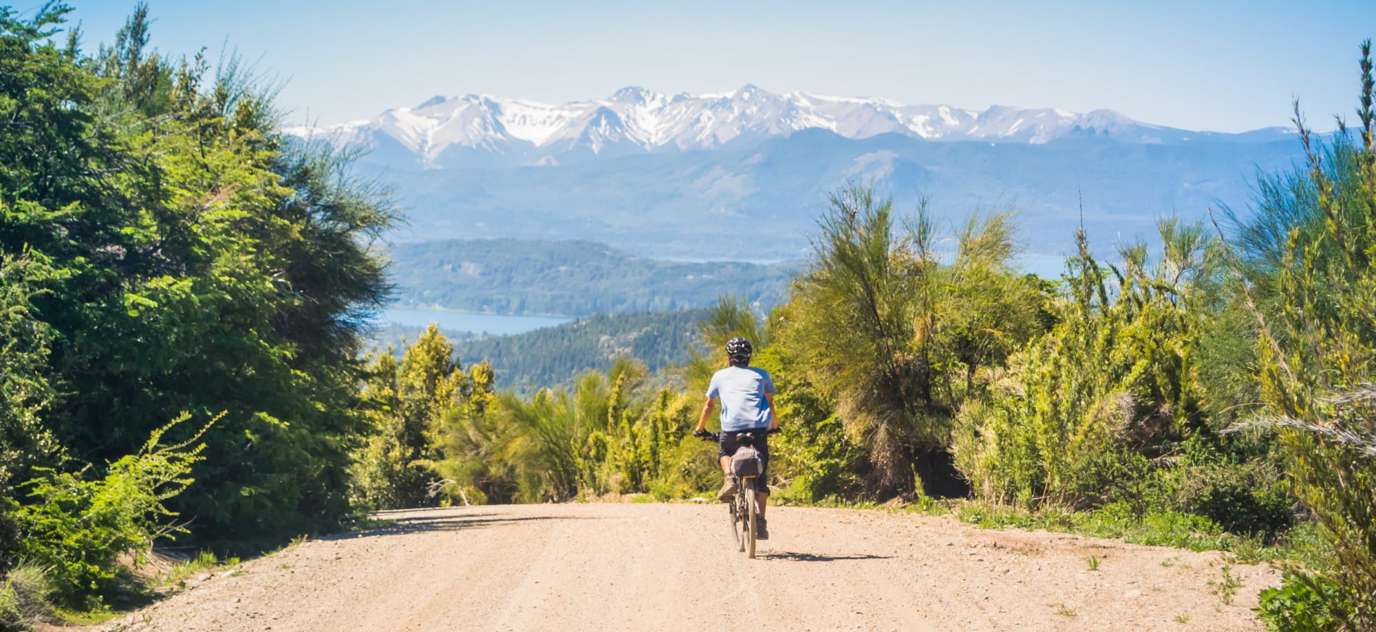 A man is cycling along a dirt road with Patagonia's snow-capped mountains in the distance.