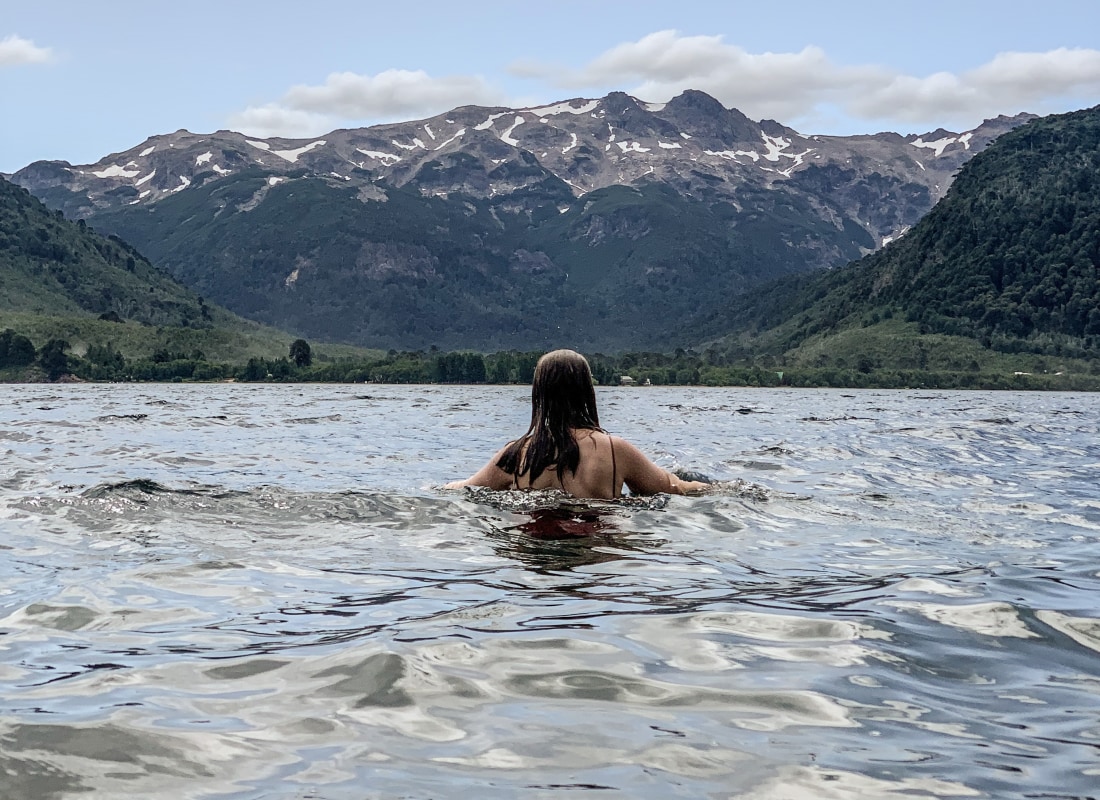 A woman is swimming in a lake alone, with snow-capped mountains in the distance. 