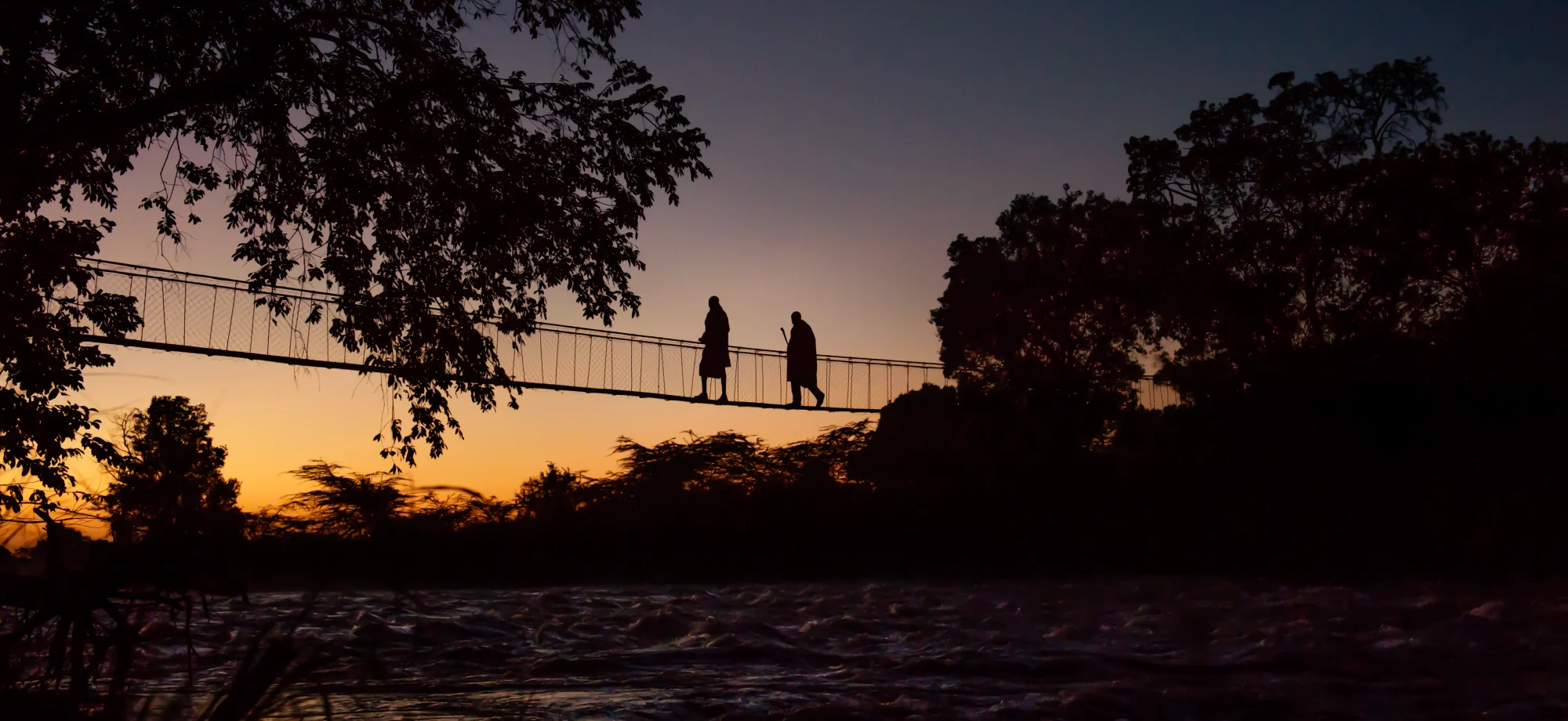 Two tribal men walk across the Ngare bridge at sunset. It is a long rope bridge joined by foliage at either side.