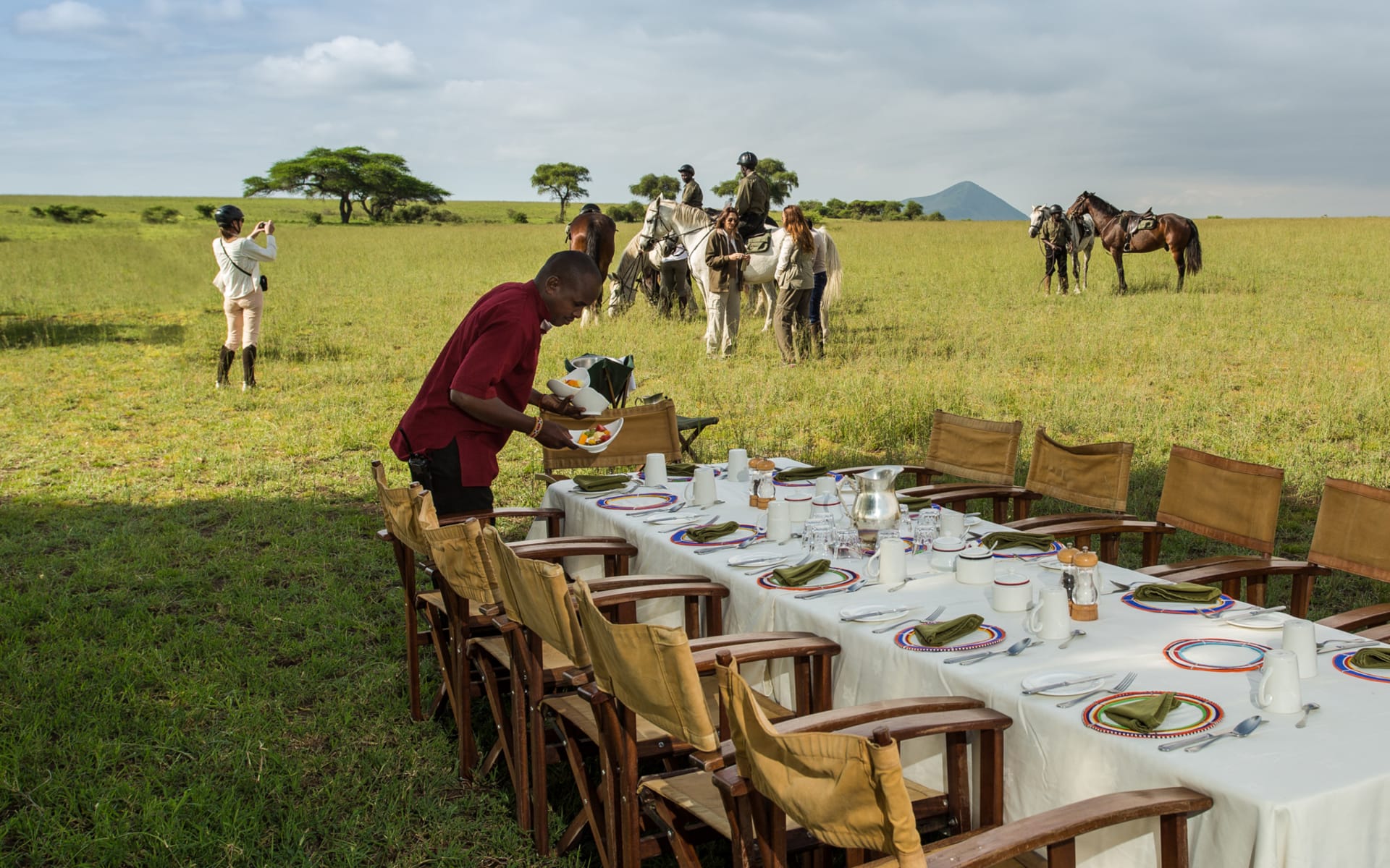 A man is setting up a long table in Amboseli National Park while others are horseriding. 