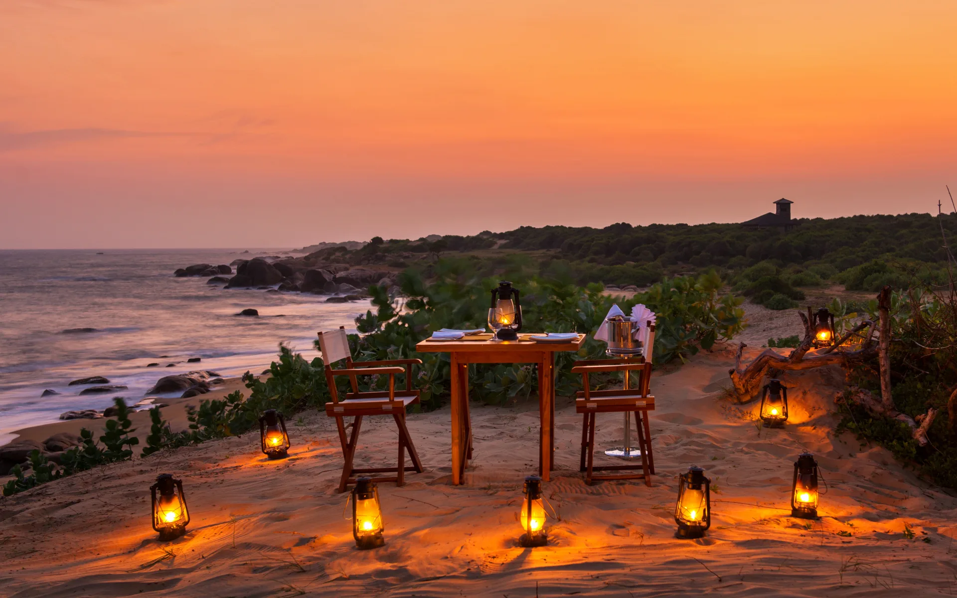 A table and chairs are surrounded by lanterns on the beach as the sun sets. 