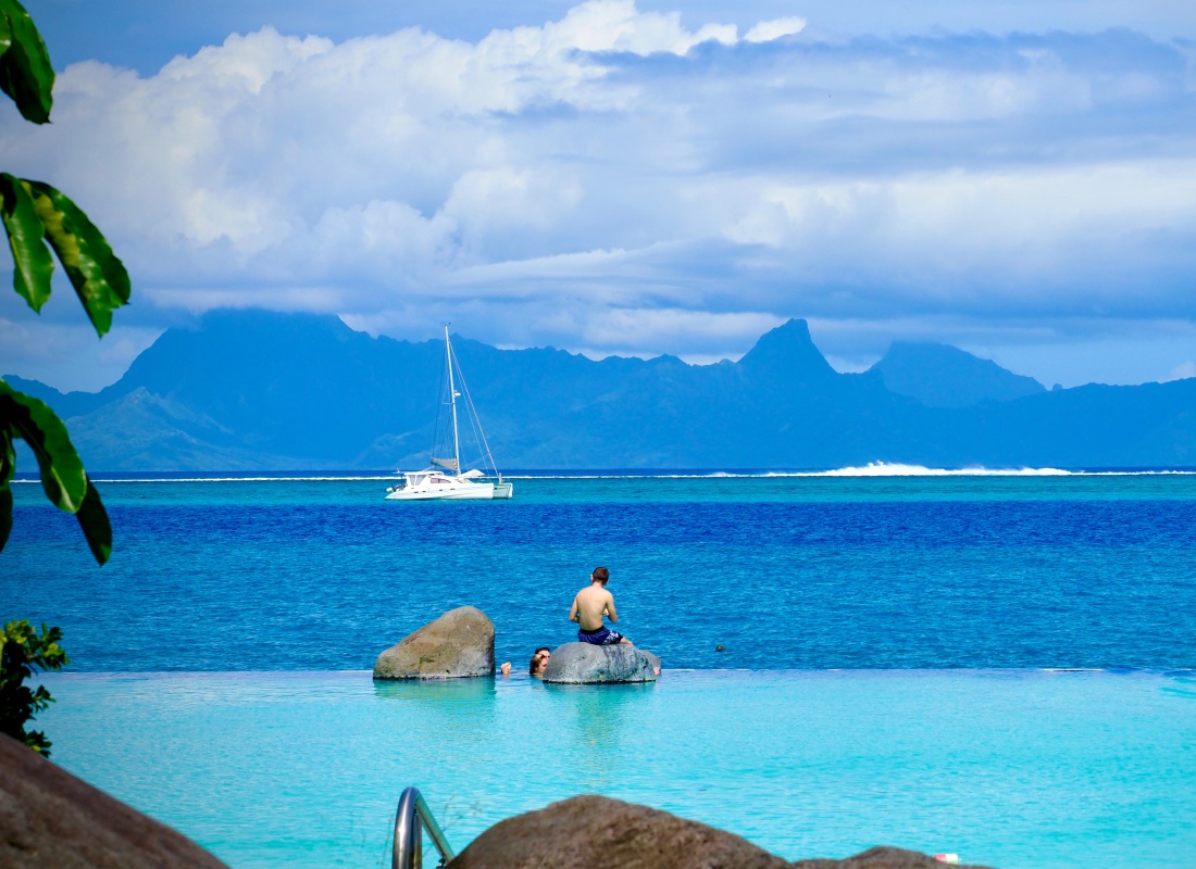 A man is sitting on a rock in the middle of an infinity pool, which overlooks the ocean and a sailing boat. 