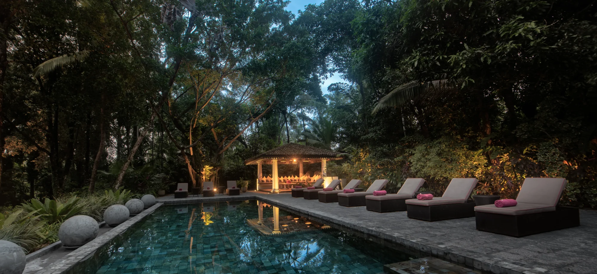 The outdoor pool is surrounded by a stone-paved sun deck equipped with comfy sun beds. The evening light shines through the thick foliage.