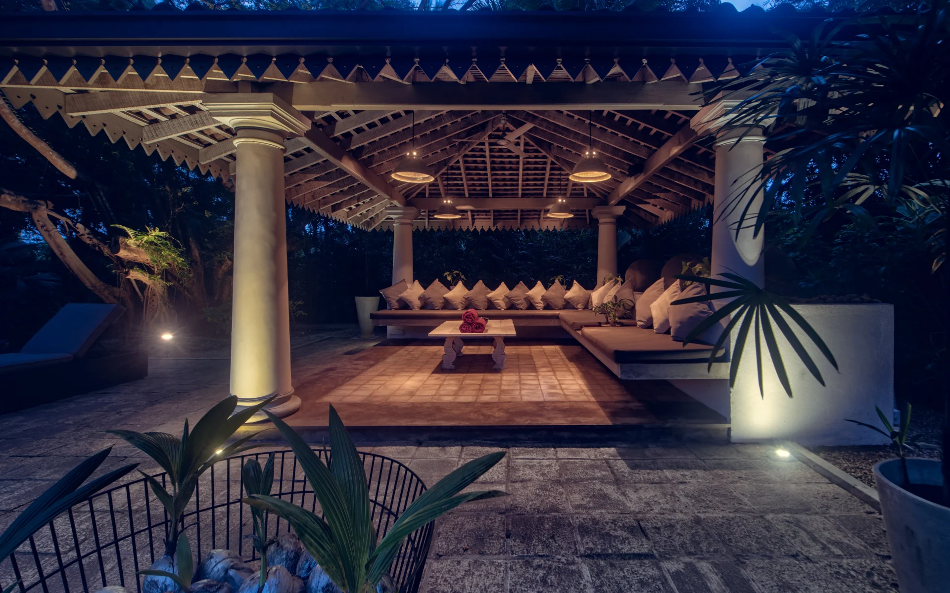 The outdoor communal living space is dimly lit during the evening. A large sofa is under a wooden shelter facing a coffee table.