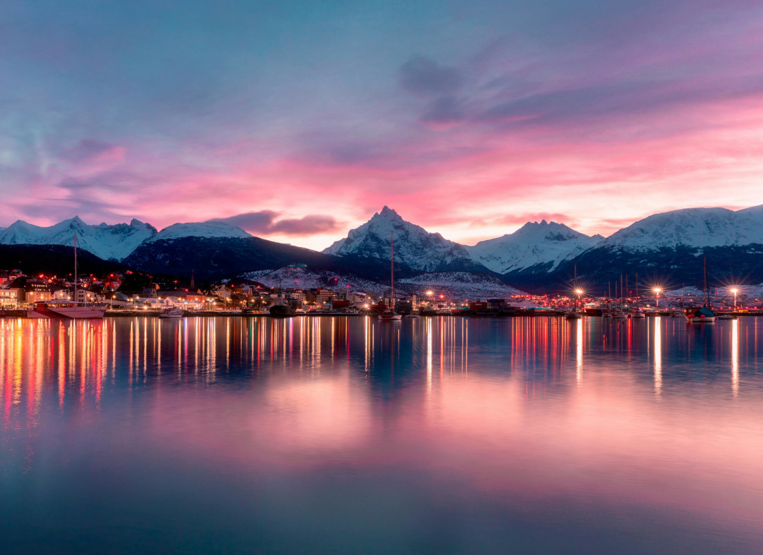 Tierra del Fuego sits on the waterfront with snow-capped mountains behind. The sky is dark, with pink and purple hues strewn across the water.