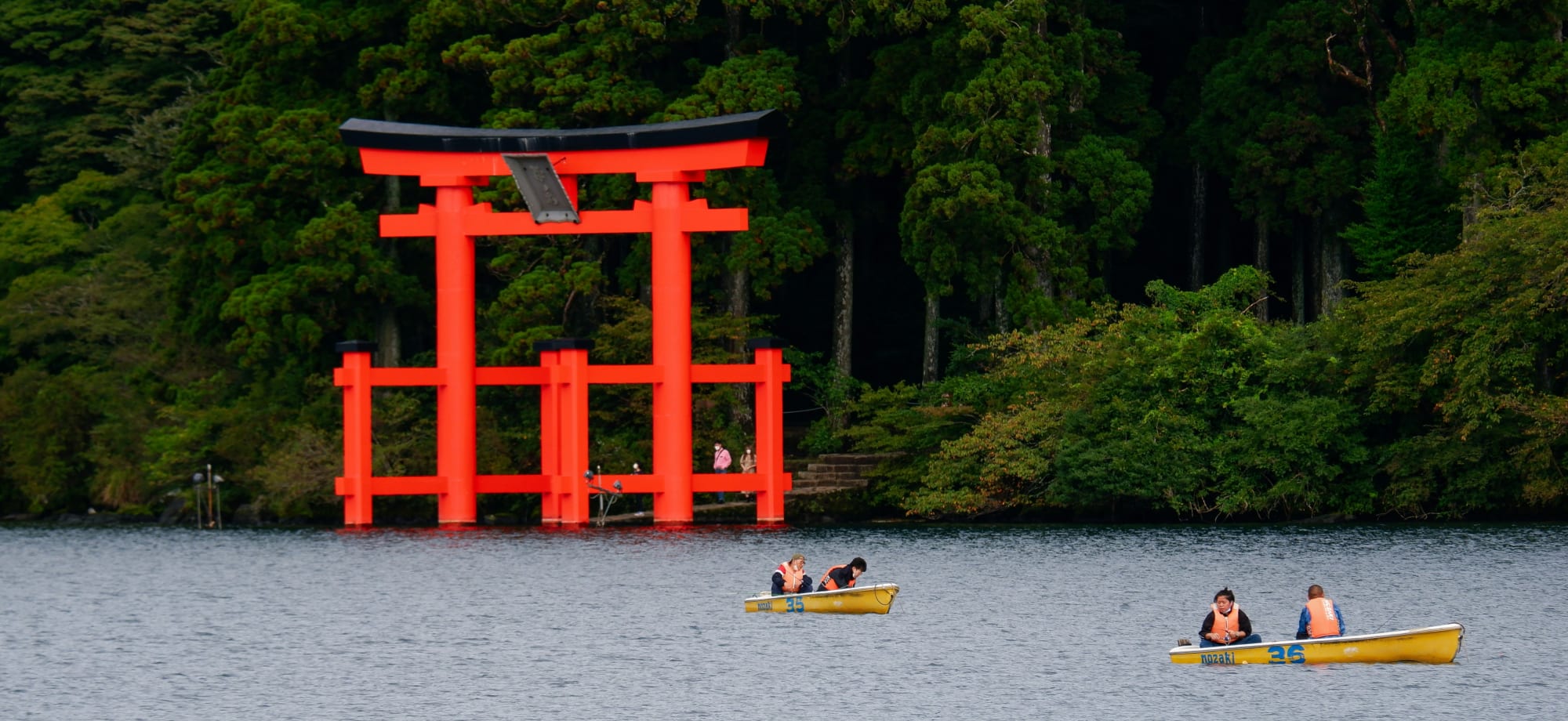 Two small boats, with two passengers each, are sailing past the bright red Torii gates in Hakone. 
