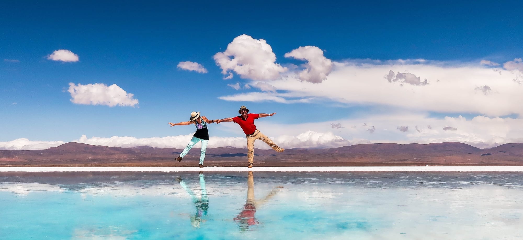 A man and woman are dancing across Salinas Grande Salt Flats' white terrain, and they are reflected in a turquoise pool. 