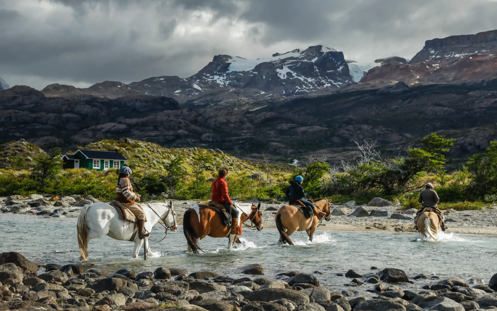 Three people ride on horses across a running stream and a dramatic mountain scenery rises behind them.