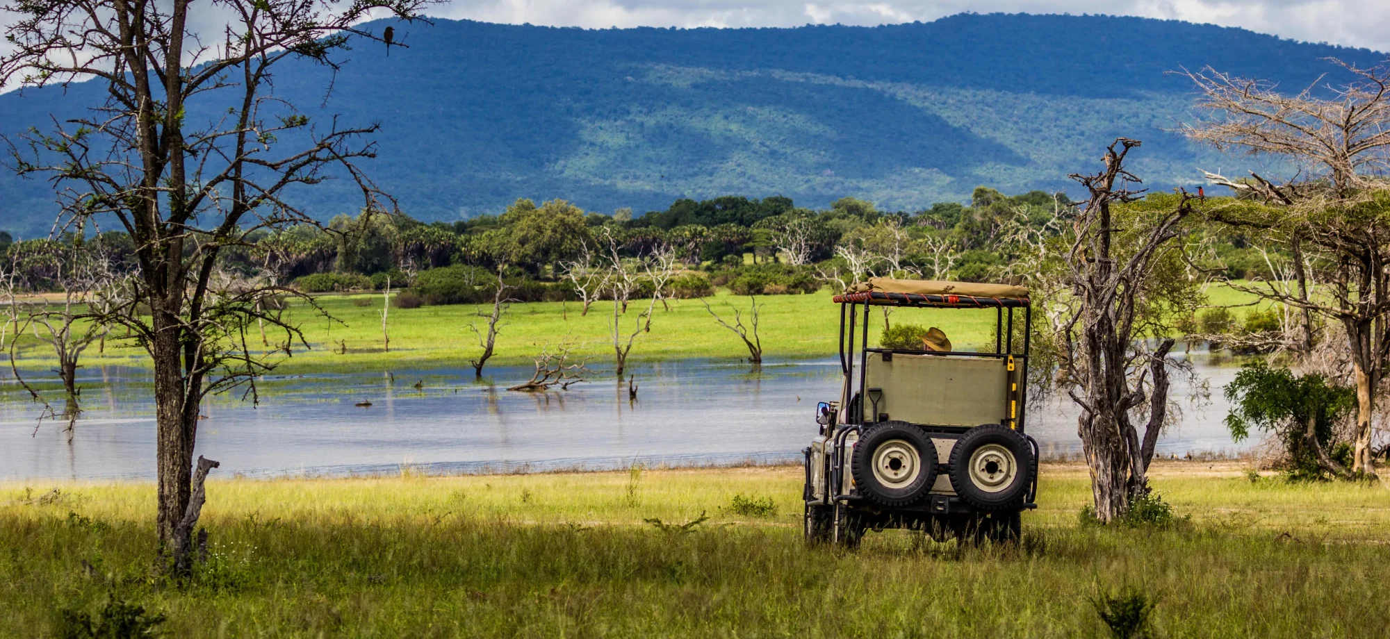A 4x4 game vehicle is parked up on the banks of the lake at Nyerere National Park, facing the tumbling, grassy mountains behind.