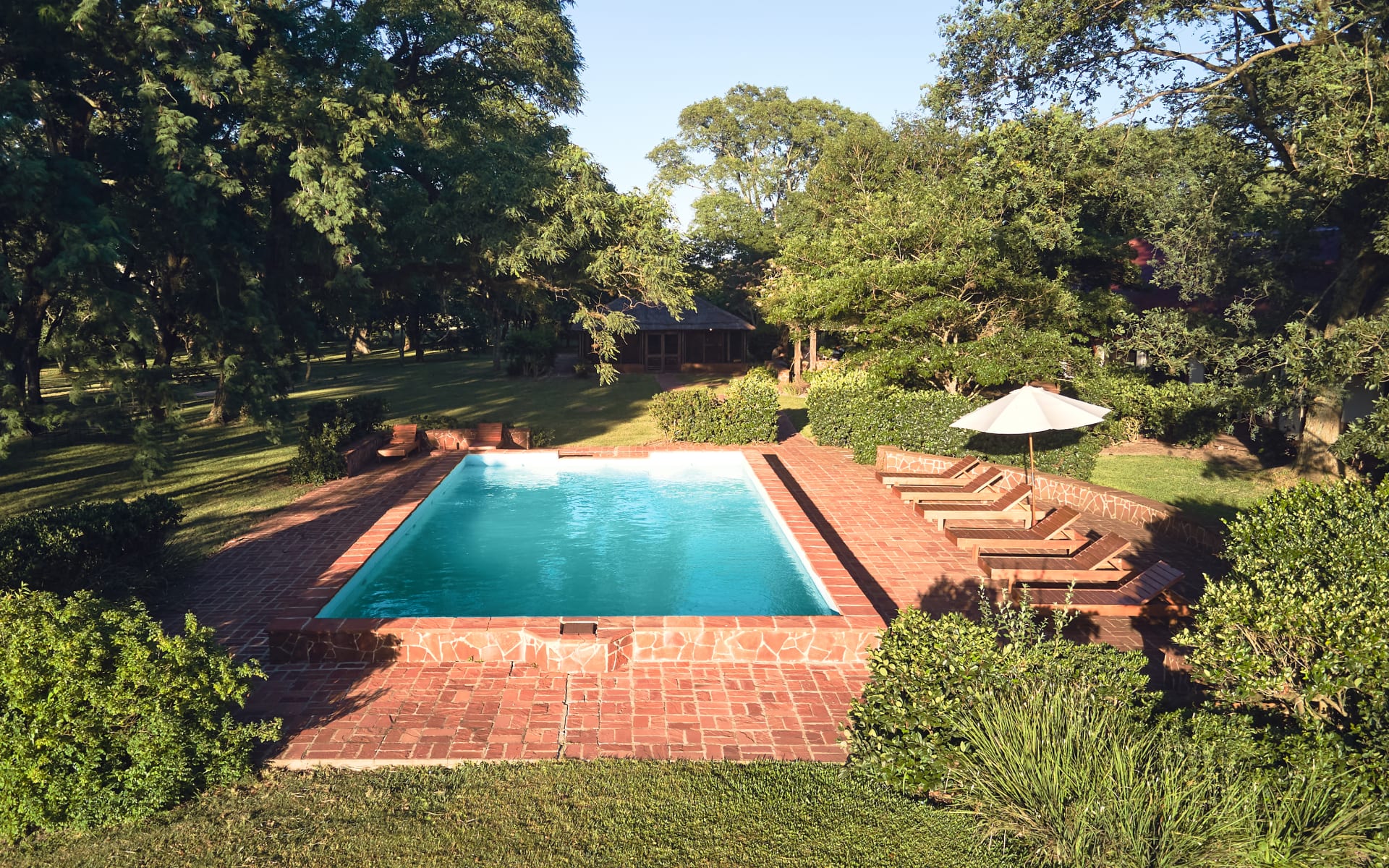 A rectangular pool sits within a red-brick deck, where sun loungers and white umbrellas stand.  