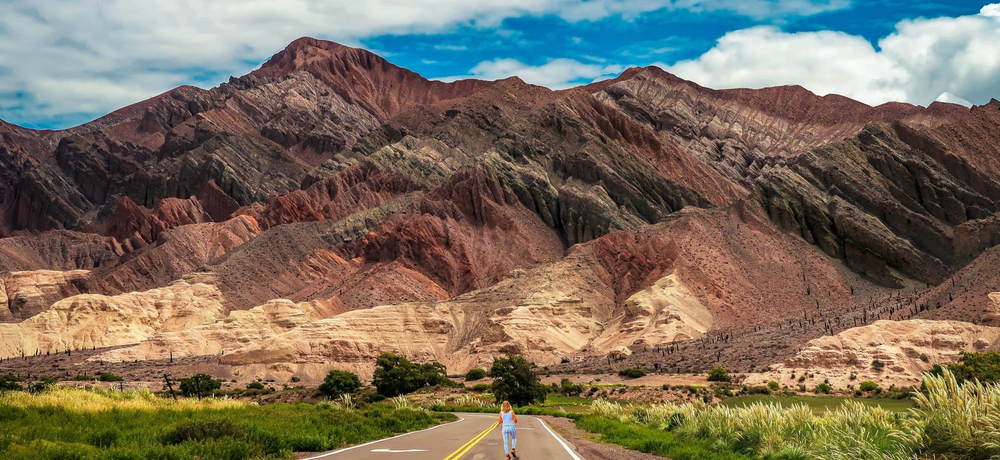 A woman is walking in the middle of a road with layers of of red-brown mountains in the background, taking up most of the sky.