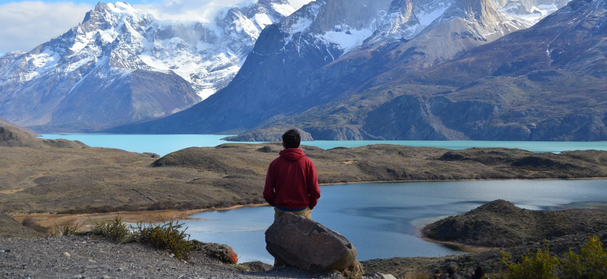 A man is standing in front of a lake and snow-capped mountains. 