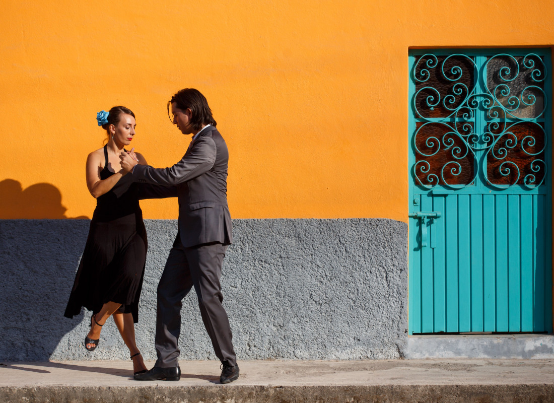 A man and woman are dancing the tango in front of a building with a yellow wall and turquoise door.