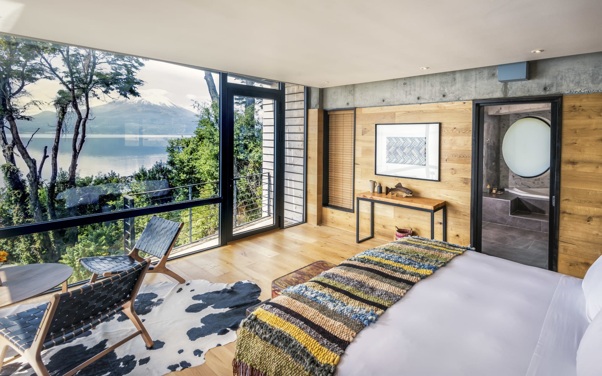 A large bed with a colourful blanket looks out of a floor-to-ceiling window onto the lake.