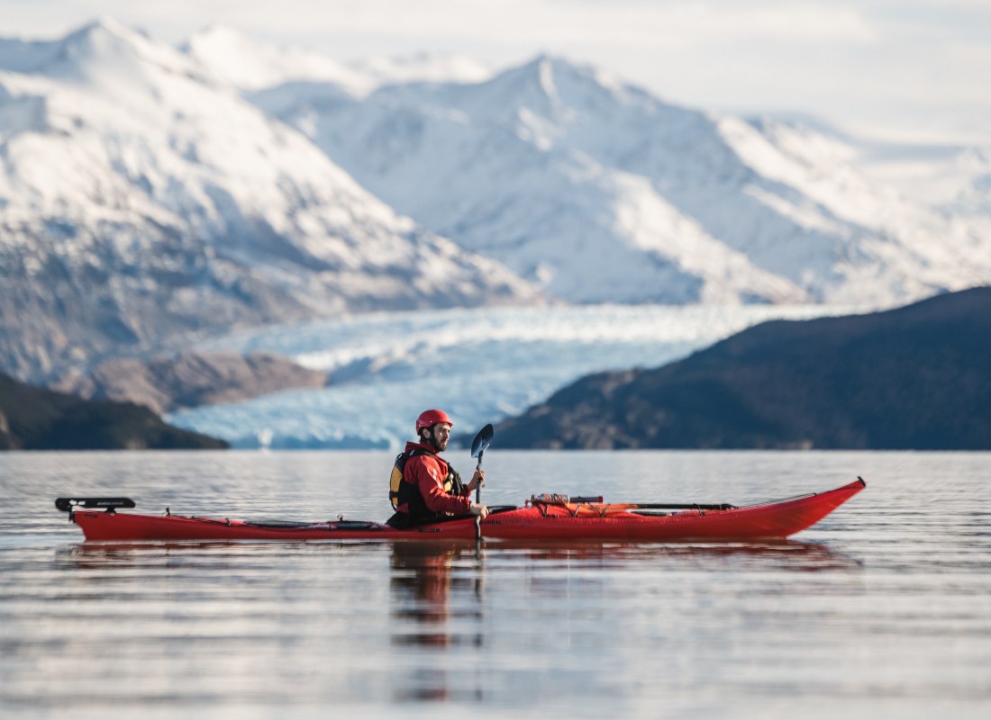 A man sits in a red kayak in the middle of the sea, backed by snowy white mountain peaks.