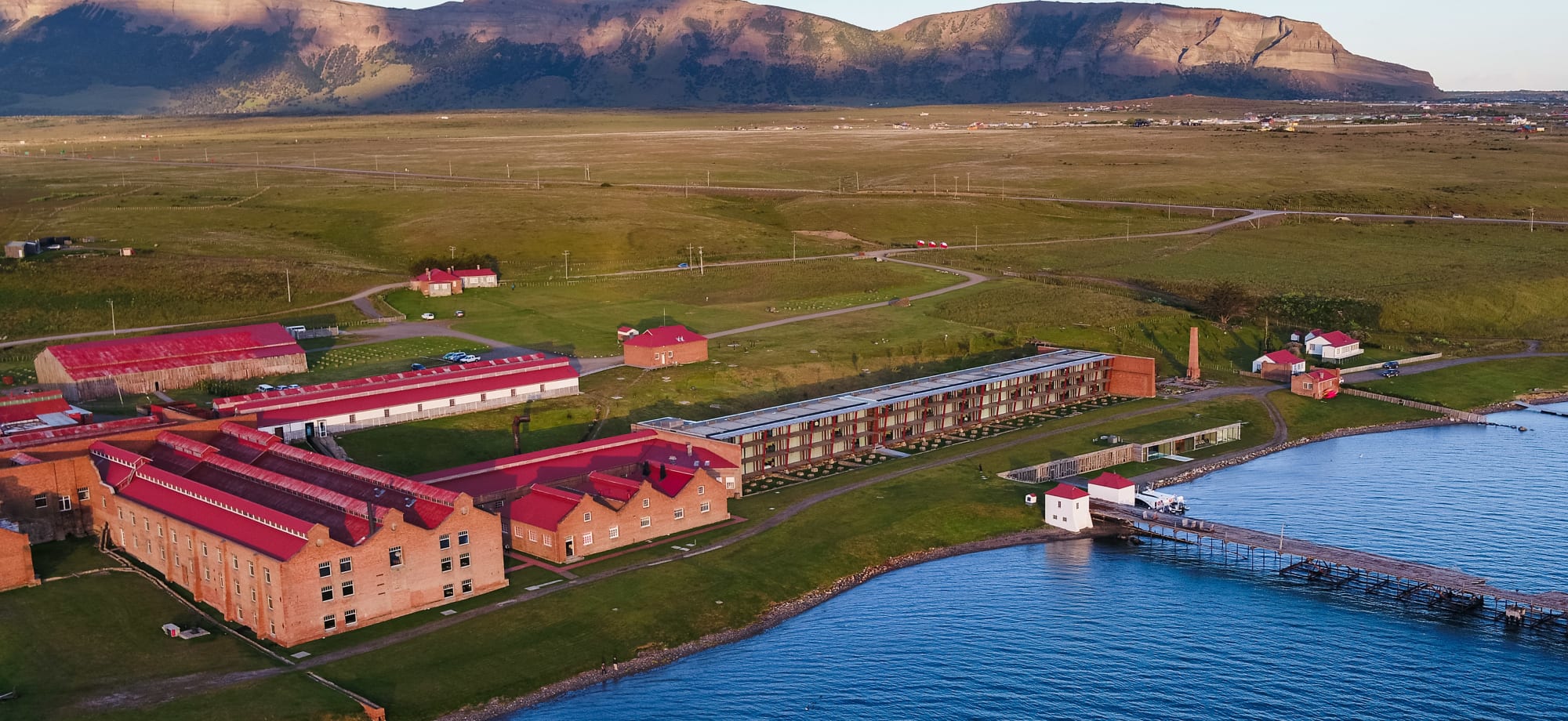 The Singular Patagonia is a long, red-roofed building on the waterfront with mountains in the background.