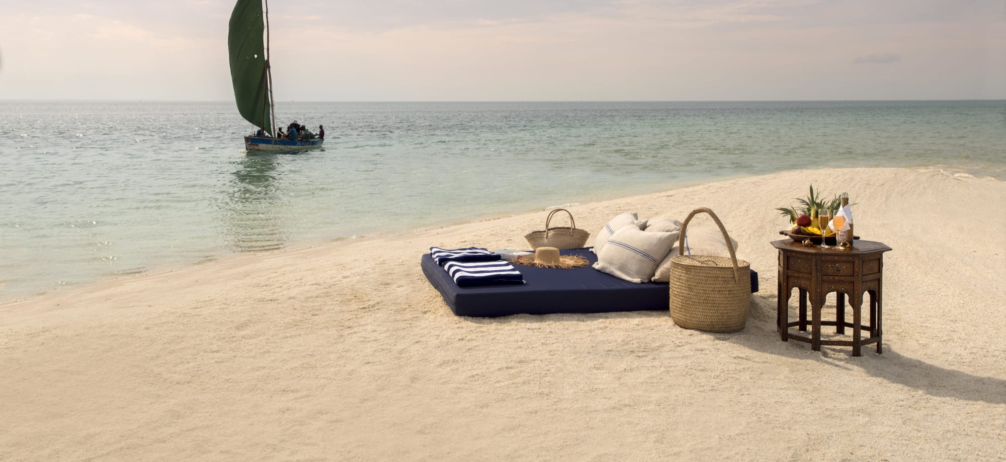 A sailing boat sits in the sea while a picnic table and blanket is laid out on the sand.
