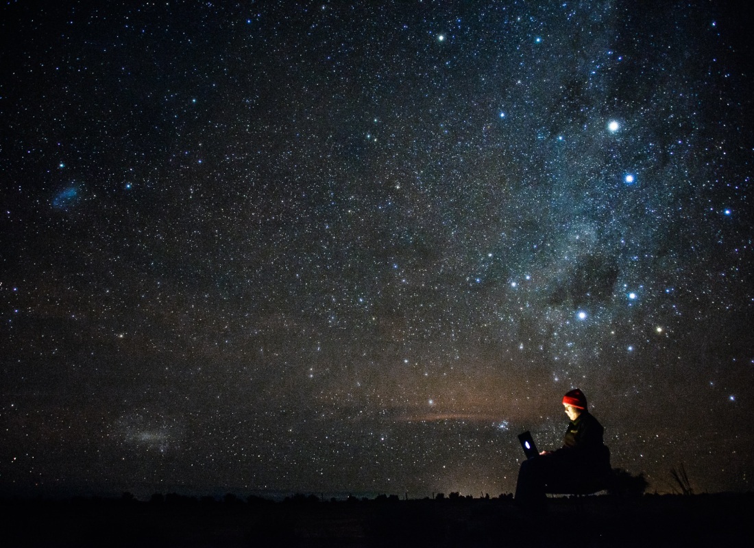 A person sits with a torch under a black sky twinkling with thousands of stars. 