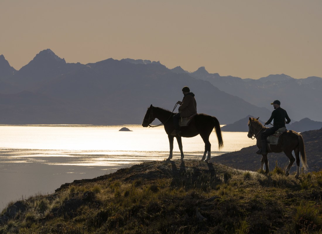 Two men are horseriding over mountains as the sun sets, cascading orange hues over a lake.