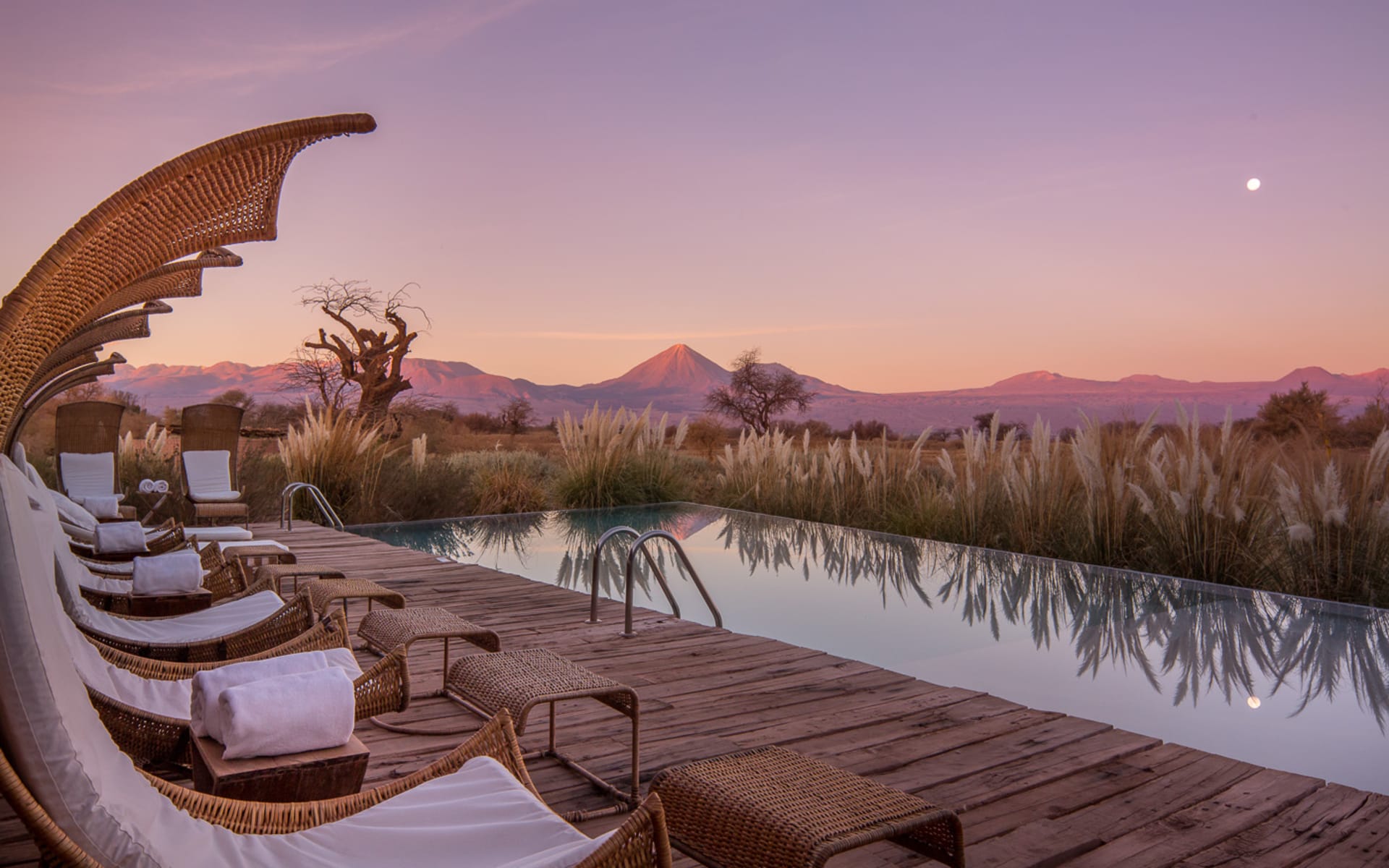 Crescent-shaped sun loungers overlook the pool and mountains in the distance that are purple from the sunset. 