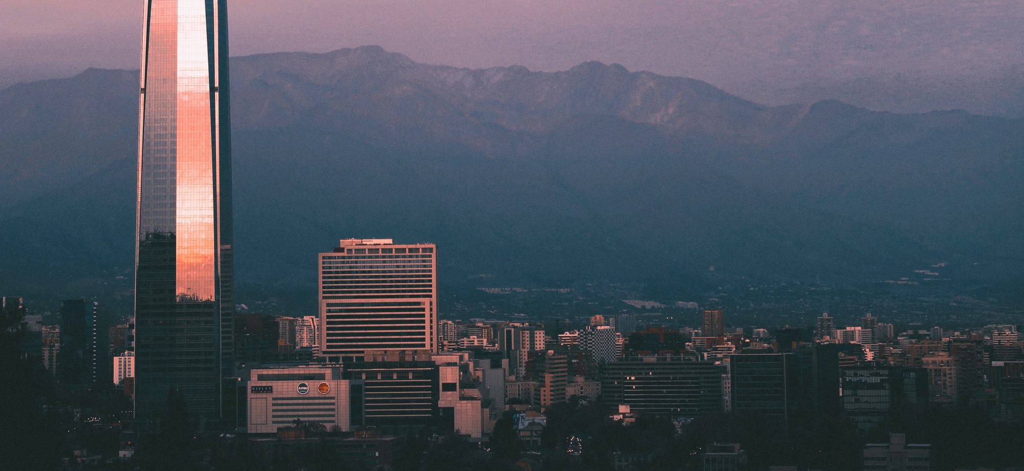 As the sun sets in Santiago, the city's skyscrapers are illuminated with an orange glow while the Andes in the background descend into darkness.