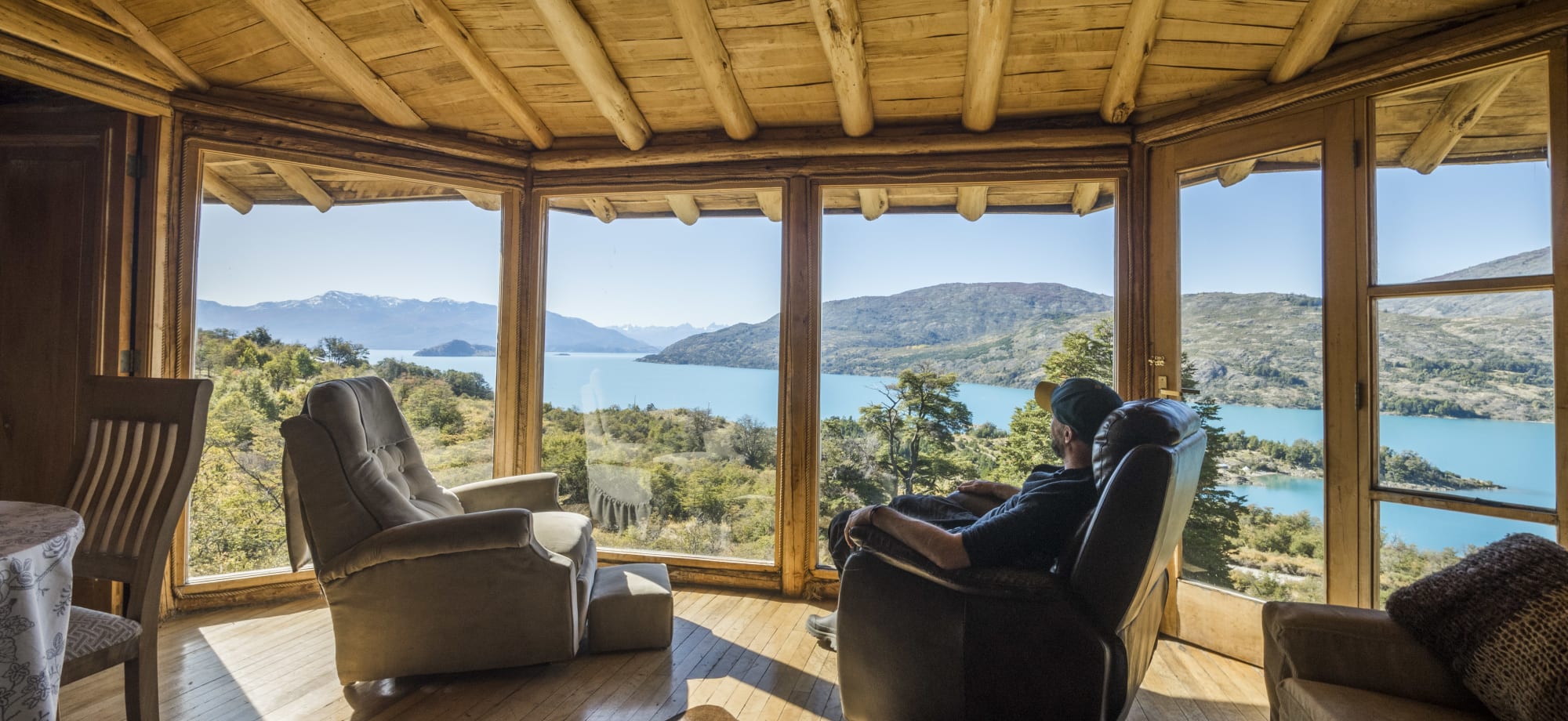 A man sits in a lounge chair looking out of a 360-degree window, offering a view of mountains and a lake.